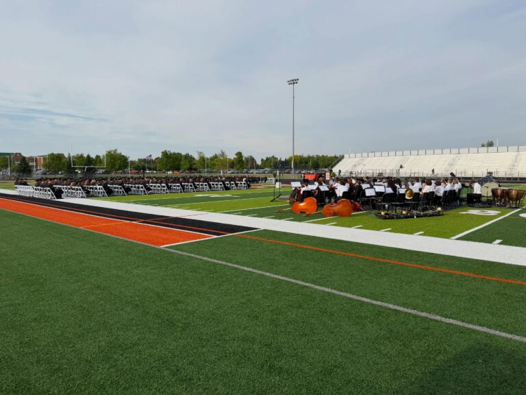 Graduates seated on a football field with families filling the stands
