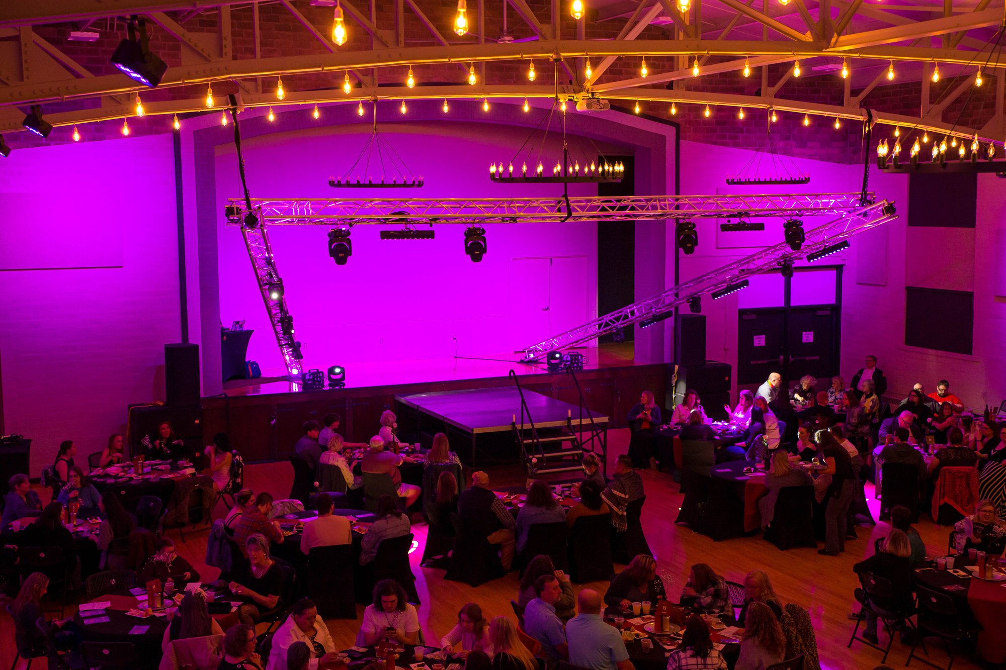 Banquet hall washed in magenta light with a truss-framed stage and guests at round tables