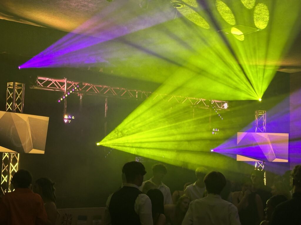 Lime green and purple light beams fan out from a truss rig over a school dance floor, with students watching from the floor.