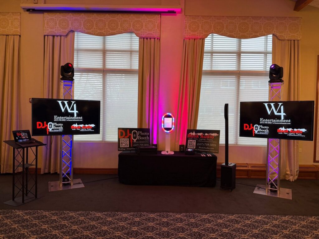 Reception setup with a DJ booth between two TVs on truss totems and warm uplighting