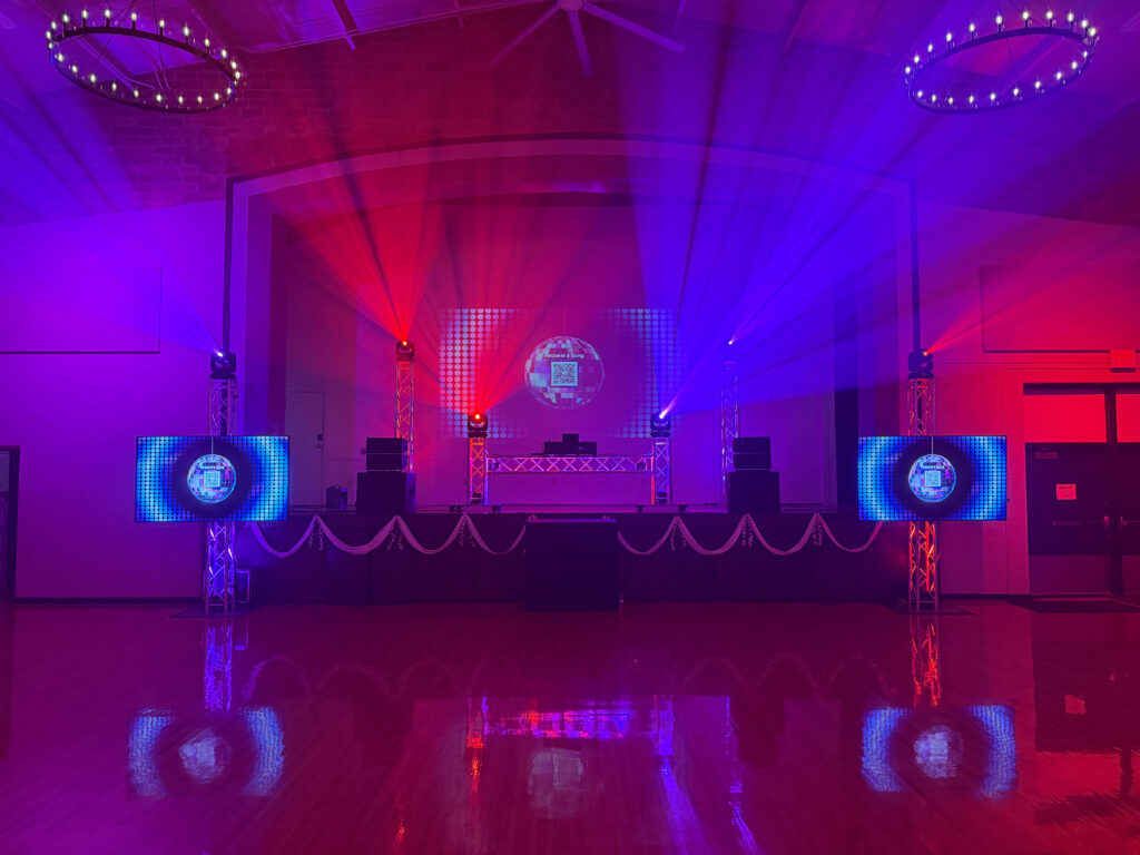The stage at the Adrian Armory lit in purple and red, with moving lights, two video screens and a DJ booth.